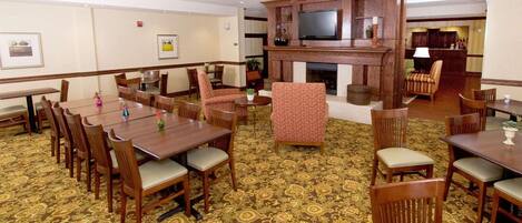 A hotel lobby with multiple dining tables and chairs, a fireplace, and a television mounted on a wooden wall.