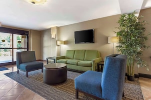 Un hall de hotel moderno con un sofá verde, sillones azules, una mesa de centro redonda, un televisor de pantalla plana y una planta grande en maceta.