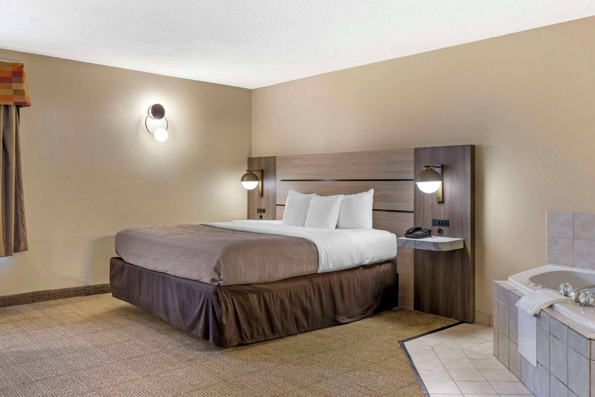 A hotel room with a large bed, a bedside table with a lamp, a built-in vanity with a mirror, and a bathtub.