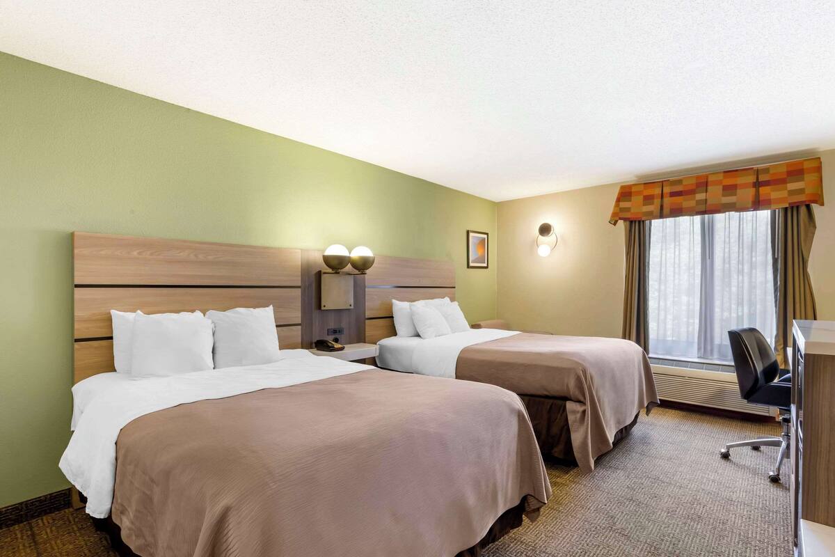 A hotel room with two beds, a desk, a chair, a window with curtains, and a wall-mounted lamp.