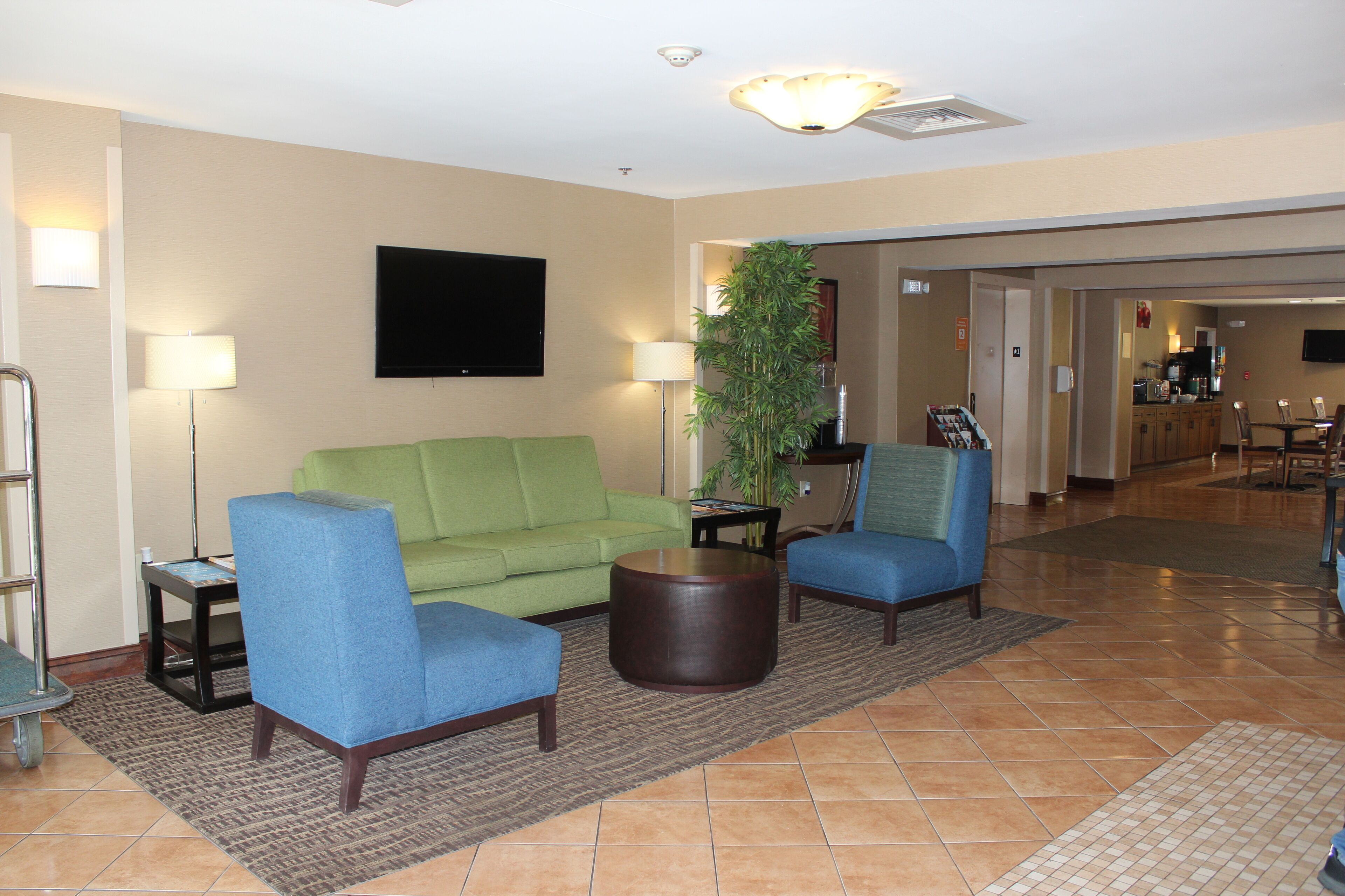Un hall d’hôtel avec un canapé vert, deux fauteuils bleus, une table basse ronde en bois, un lampadaire et un téléviseur fixé au mur.