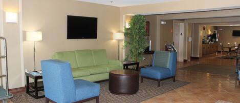 A hotel lobby with a green sofa, two blue armchairs, a round wooden coffee table, a floor lamp, and a television mounted on the wall.