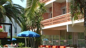 A hotel with a two-story balcony, outdoor seating area with tables and chairs, and lush greenery including palm trees. - Hotel Marina (Massa)