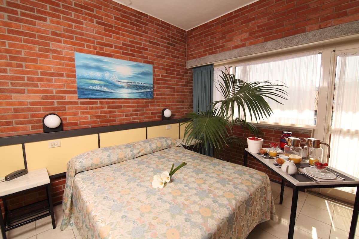 Une pièce avec un mur en briques, un lit recouvert d’une literie à motifs floraux, une table avec des éléments pour le petit-déjeuner et une grande fenêtre avec des rideaux.