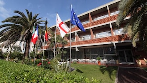 A building with multiple flags, including the EU flag, and a well-maintained garden with palm trees. - Hotel Marina (Massa)