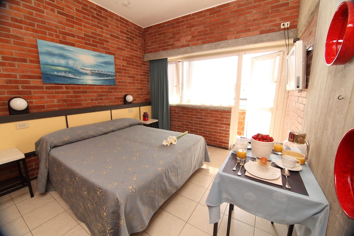 Une chambre d’hôtel avec un lit, une table à manger avec des aliments pour le petit-déjeuner, un tableau au mur et une fenêtre avec des rideaux.