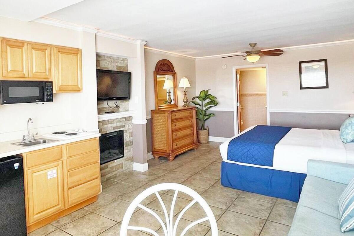 Een kleine hotelkamer met een kitchenette, een bed met blauw beddengoed, een plafondventilator en een houten dressoir.