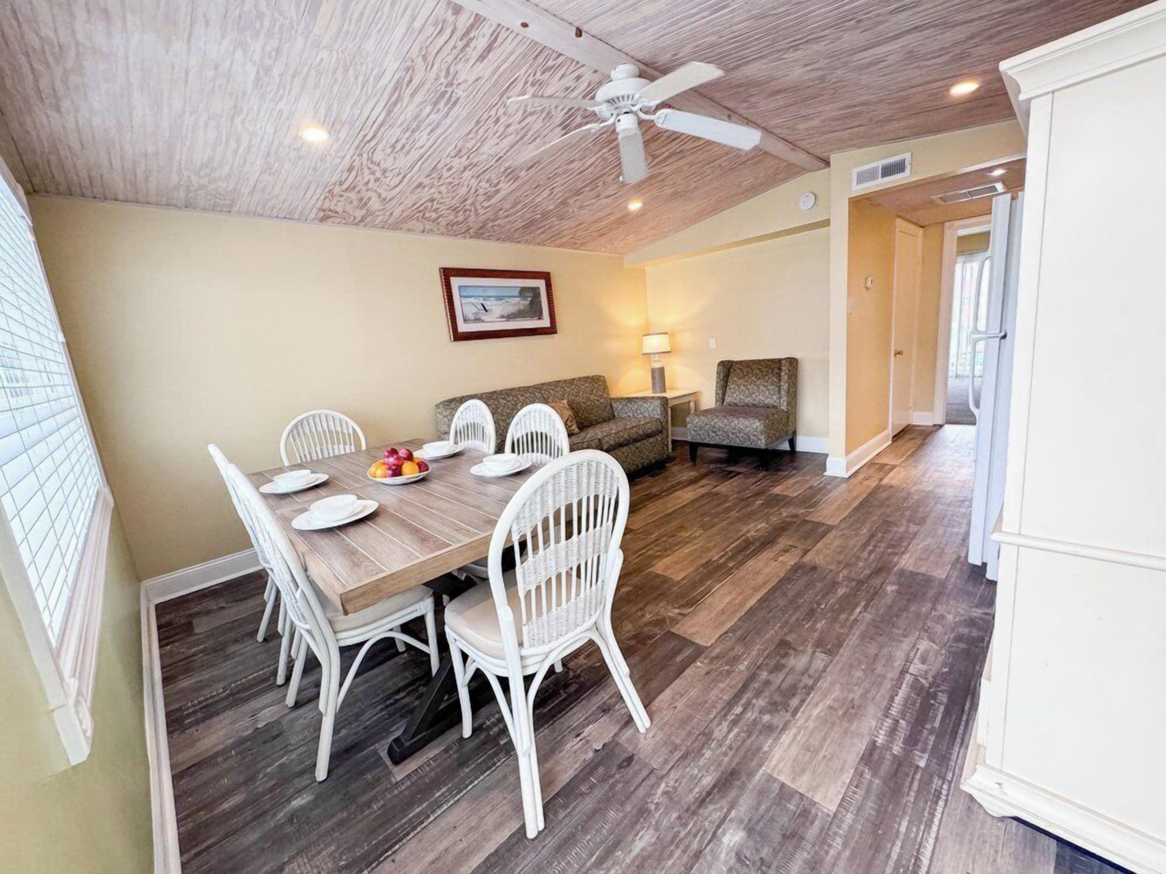 A dining area with a wooden table and white chairs, a ceiling fan, and a framed picture on the wall.