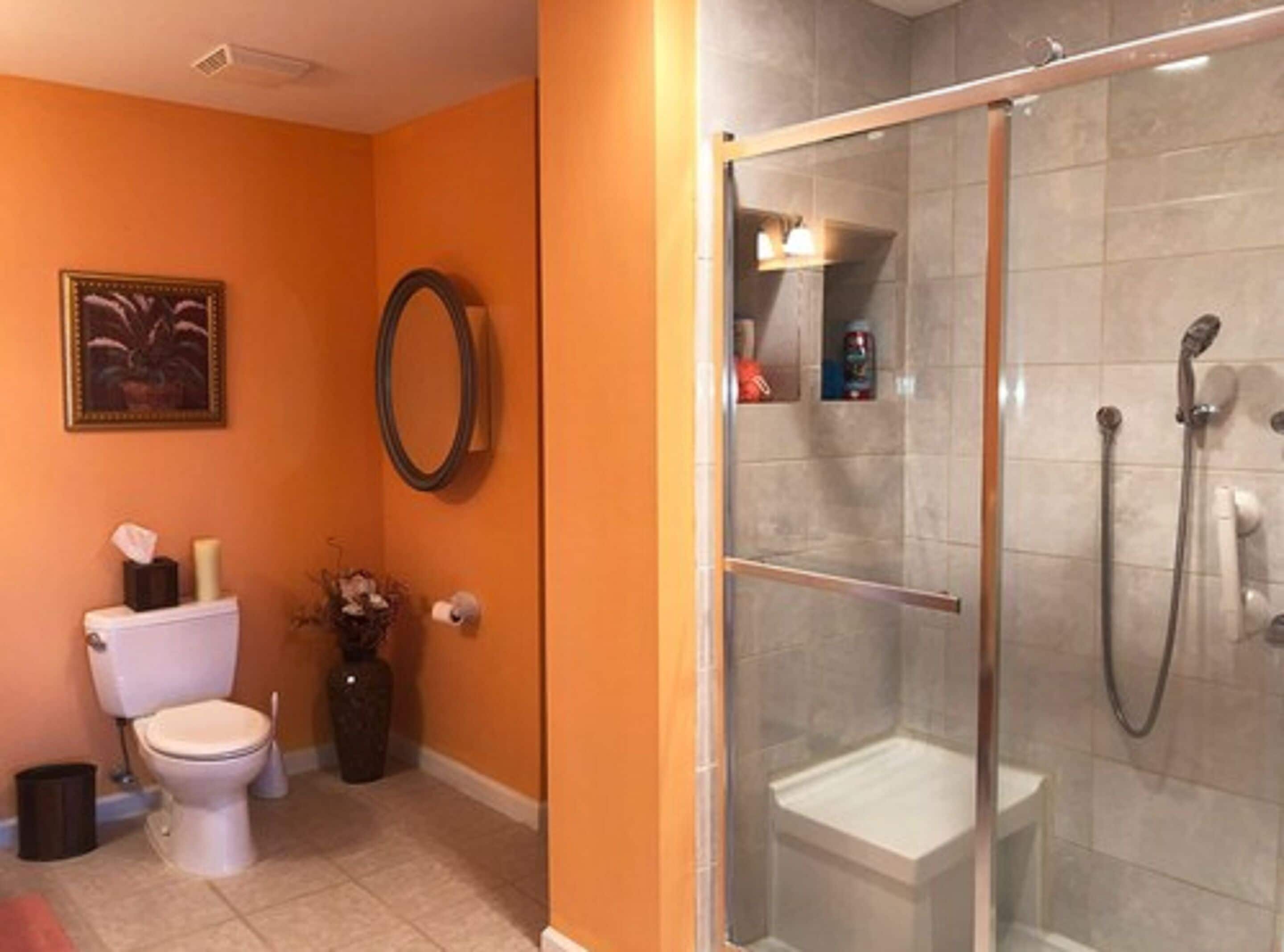 A bathroom with an orange wall, a toilet, a shower with a glass door, and a mirror.