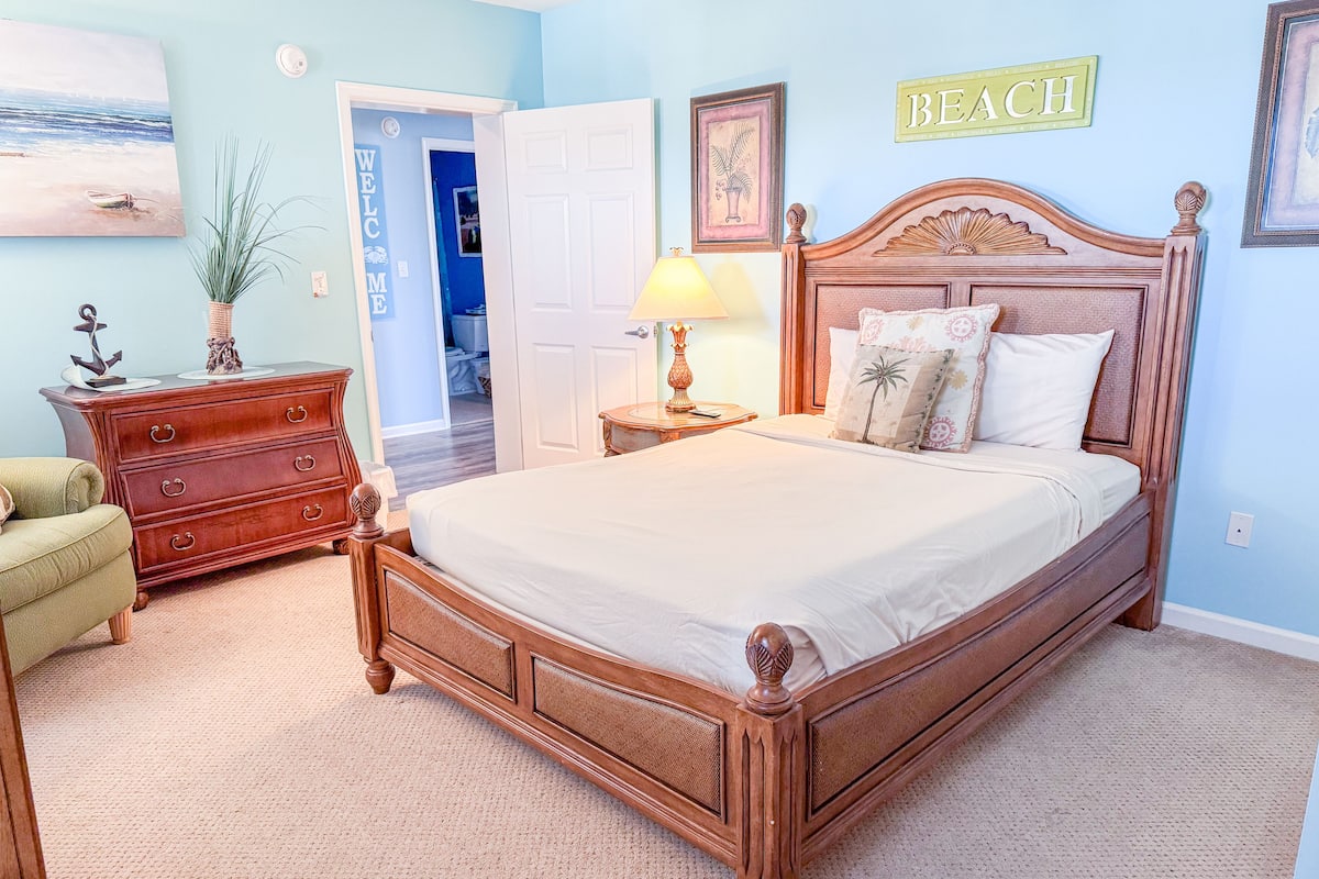 Een slaapkamer met een houten bed, een dressoir, een nachtkastje, een lamp en een deur naar een andere kamer.