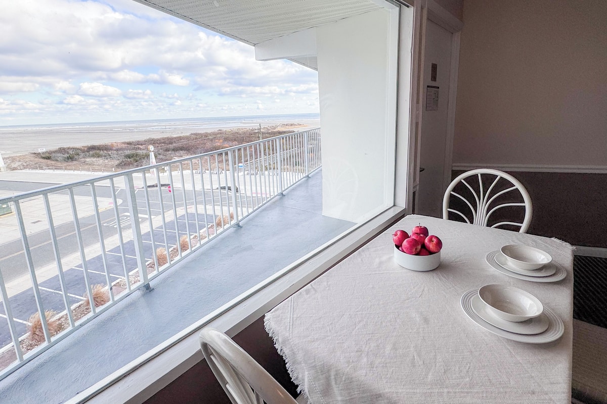 Een balkon met een tafel gedekt voor twee personen, een witte stoel en uitzicht op het strand en de oceaan.