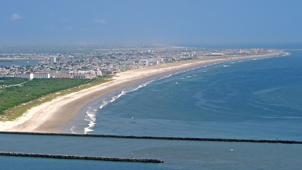 Vue aérienne d’une ville côtière avec une longue plage, un port de plaisance et un grand complexe hôtelier.