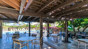 An outdoor patio area with tables and chairs, a barbecue grill, and a pool in the background. - Maui Kamaole (Kihei)