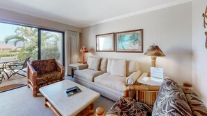 A living room with a sofa, armchair, coffee table, and a desk with a lamp. There are two framed pictures on the wall, and a window with a view of palm trees. - Maui Kamaole (Kihei)