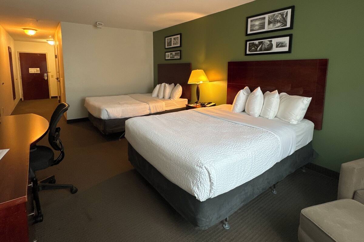 A hotel room with two beds, a desk, a chair, and a lamp.