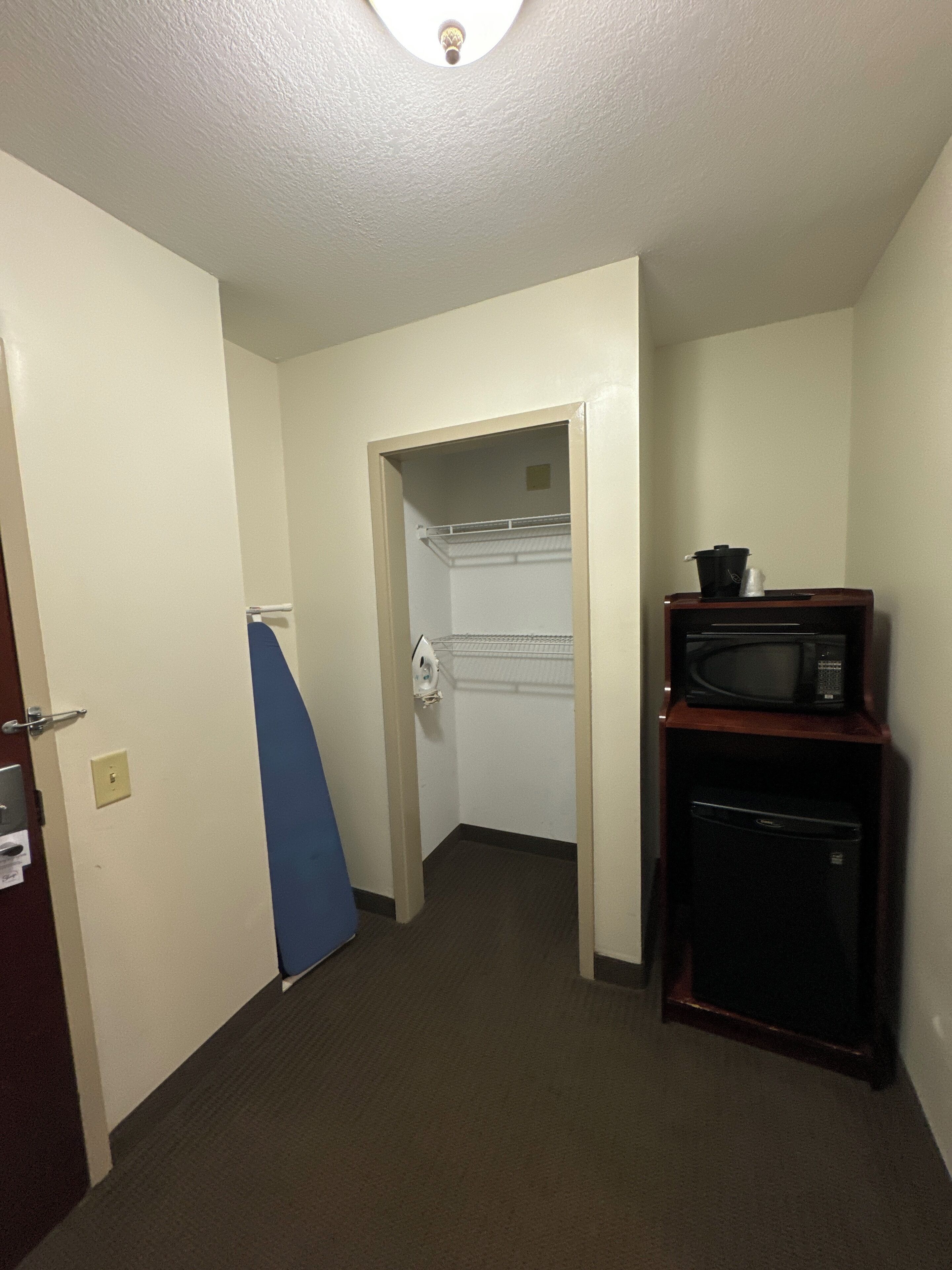 A narrow hallway with a microwave on a wooden stand, a blue ironing board, and a white door leading to a closet.
