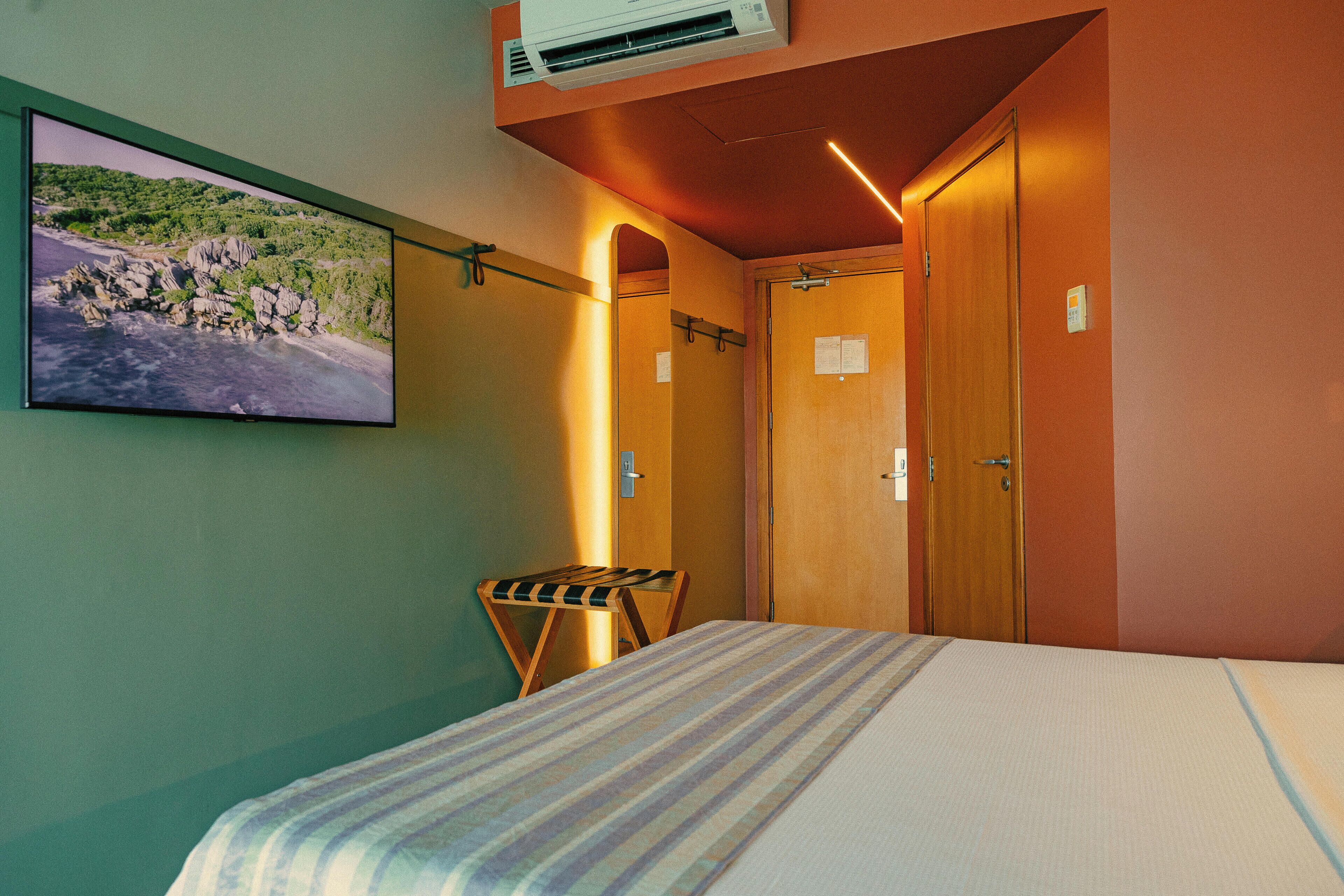 Habitación de hotel con cama, televisión montada mostrando un paisaje pintoresco, banca de madera y una puerta con un letrero.