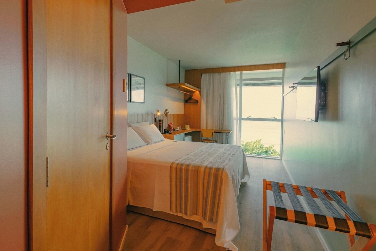 Un dormitorio con cama, mesita de noche, silla y ventana con cortinas.