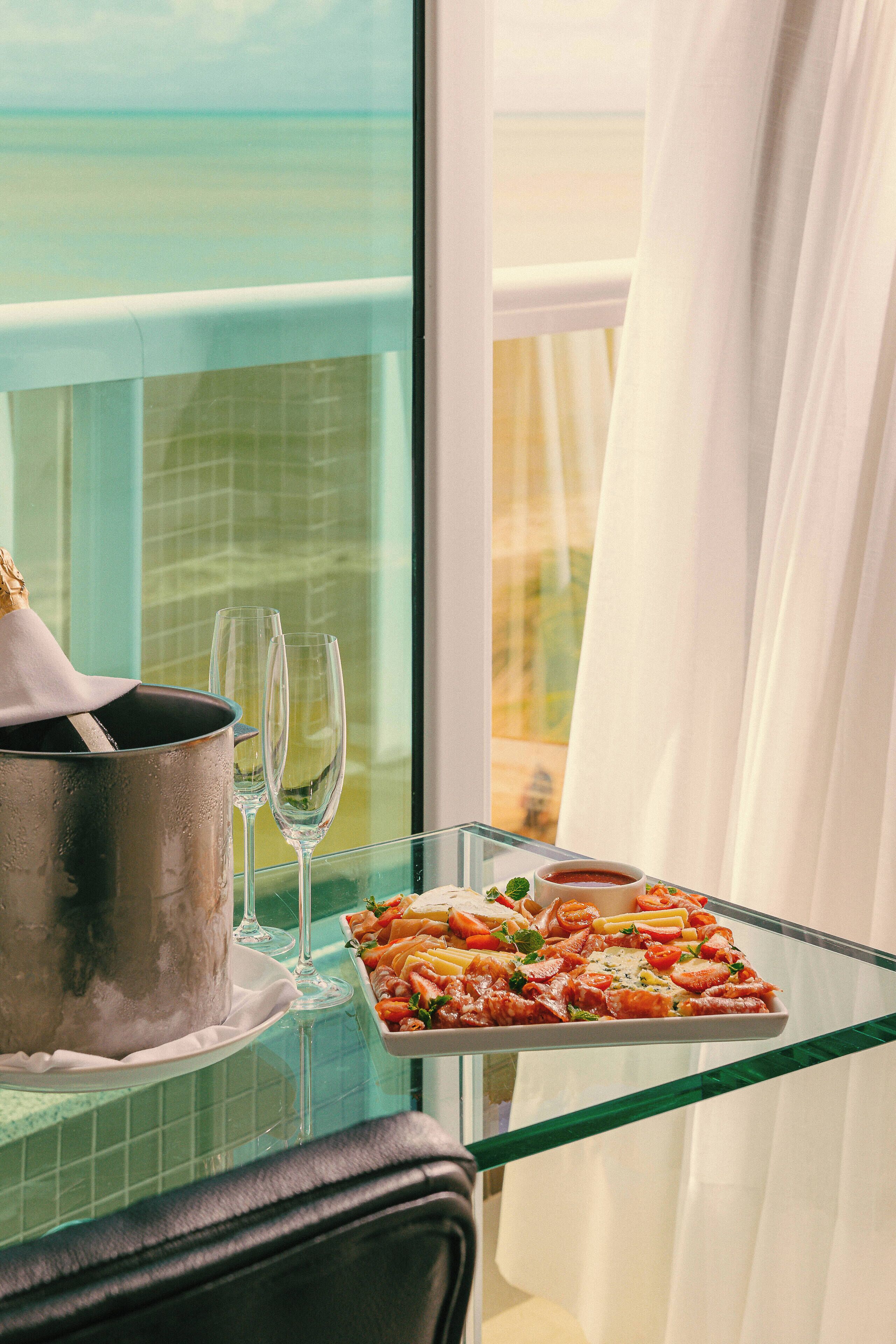 Een glazen tafel met een dienblad eten en twee wijnglazen, een champagnekoeler, en uitzicht op zee door een raam met gordijnen.