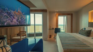 Habitación de hotel con una televisión grande, una cama, un escritorio y vista al océano.