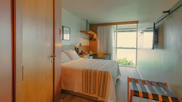 Quarto de hotel com cama, uma pequena mesa, uma cadeira, escrivaninha e janela com vista.