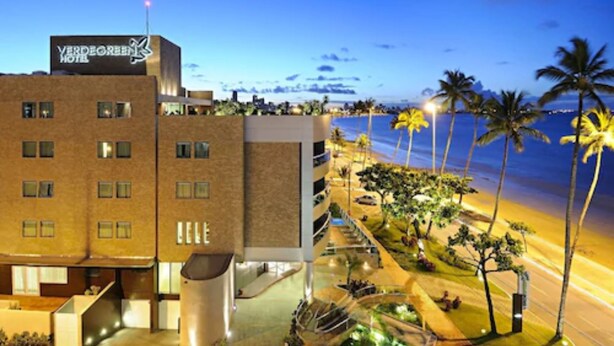 Un hotel con diseño moderno, un letrero que dice "VERDE GREEN HOTEL" y vista a la playa con palmeras y un camino que lleva a la orilla.