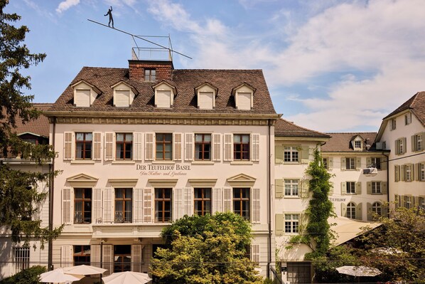 A historic building with a sign reading "Der Teufelhof Basel," featuring a classic European architectural style with symmetrical windows and a tiled roof.