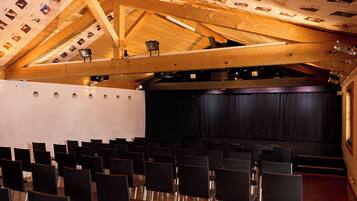 Un auditorio con hileras de sillas, un escenario con cortina y vigas de madera en el techo.