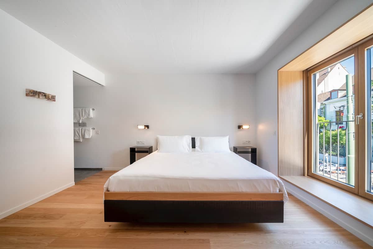 Un dormitorio moderno con una cama grande, suelo de madera, paredes blancas y una ventana con vistas al exterior.