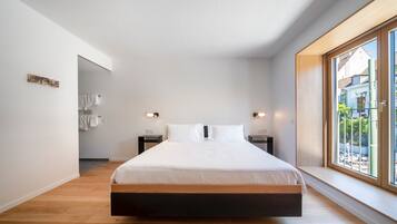 A modern bedroom with a large bed, wooden floor, white walls, and a window with a view of the outside.