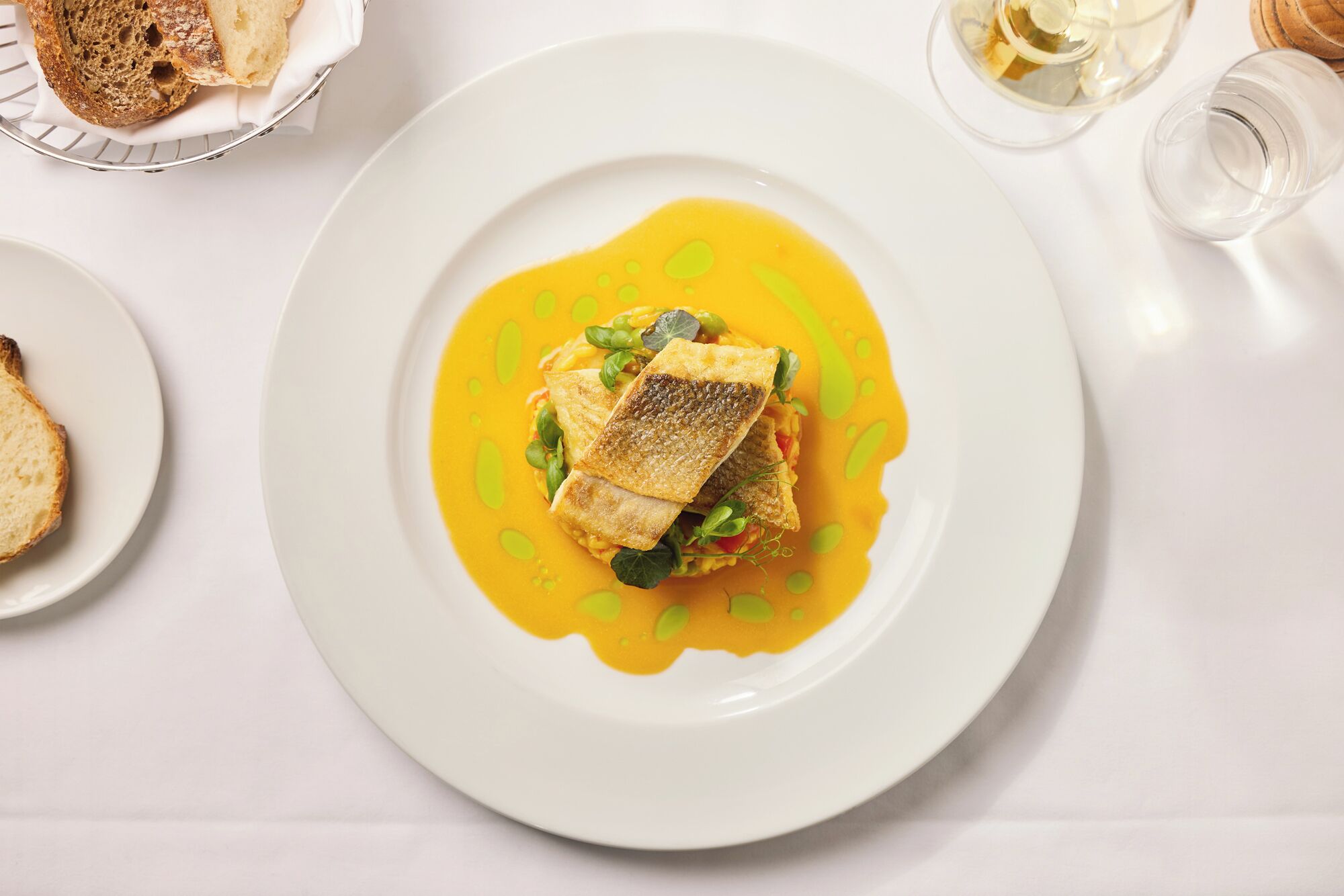 A plate of food with a fish fillet and vegetables in a yellow sauce, accompanied by slices of bread and a glass of white wine on a white table.