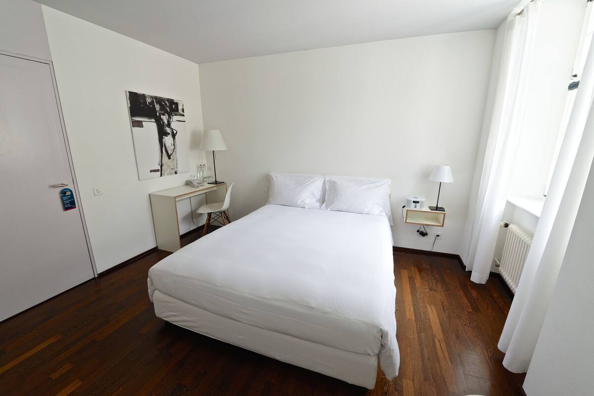 Una habitación de hotel con una cama grande, un escritorio, dos lámparas, un radiador y una ventana con cortinas.
