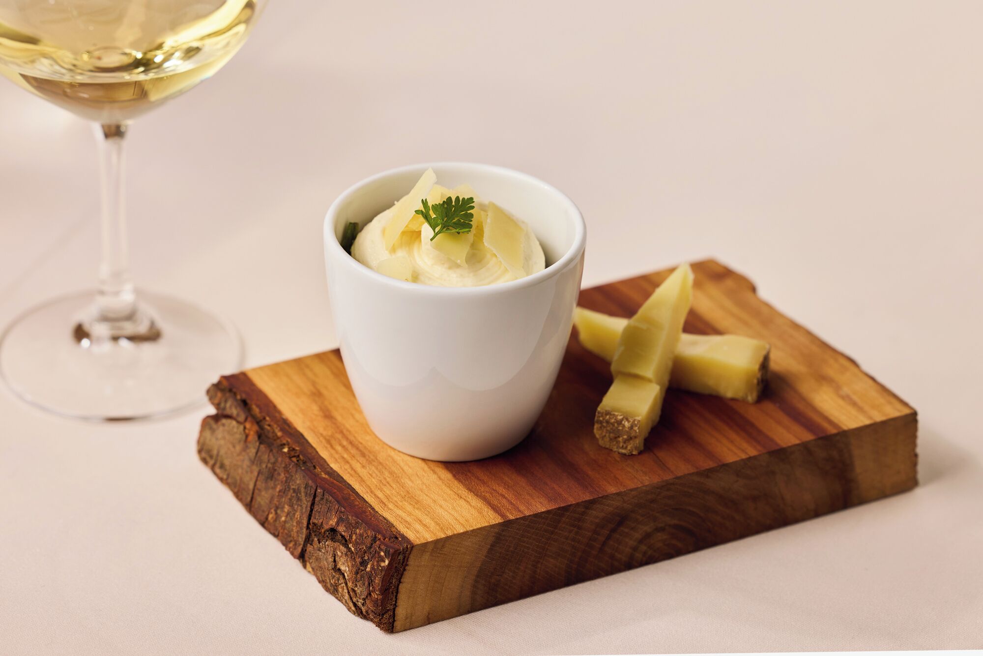 Un tazón blanco con un postre cremoso, una copa de vino blanco y una tabla de madera con lonchas de queso.
