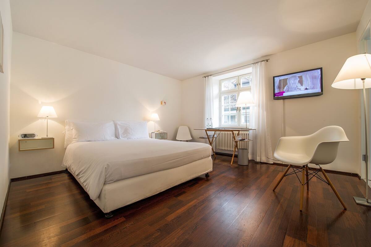 Habitación de hotel con cama, lámparas de noche, un escritorio con silla, televisor en la pared y ventana con cortinas.