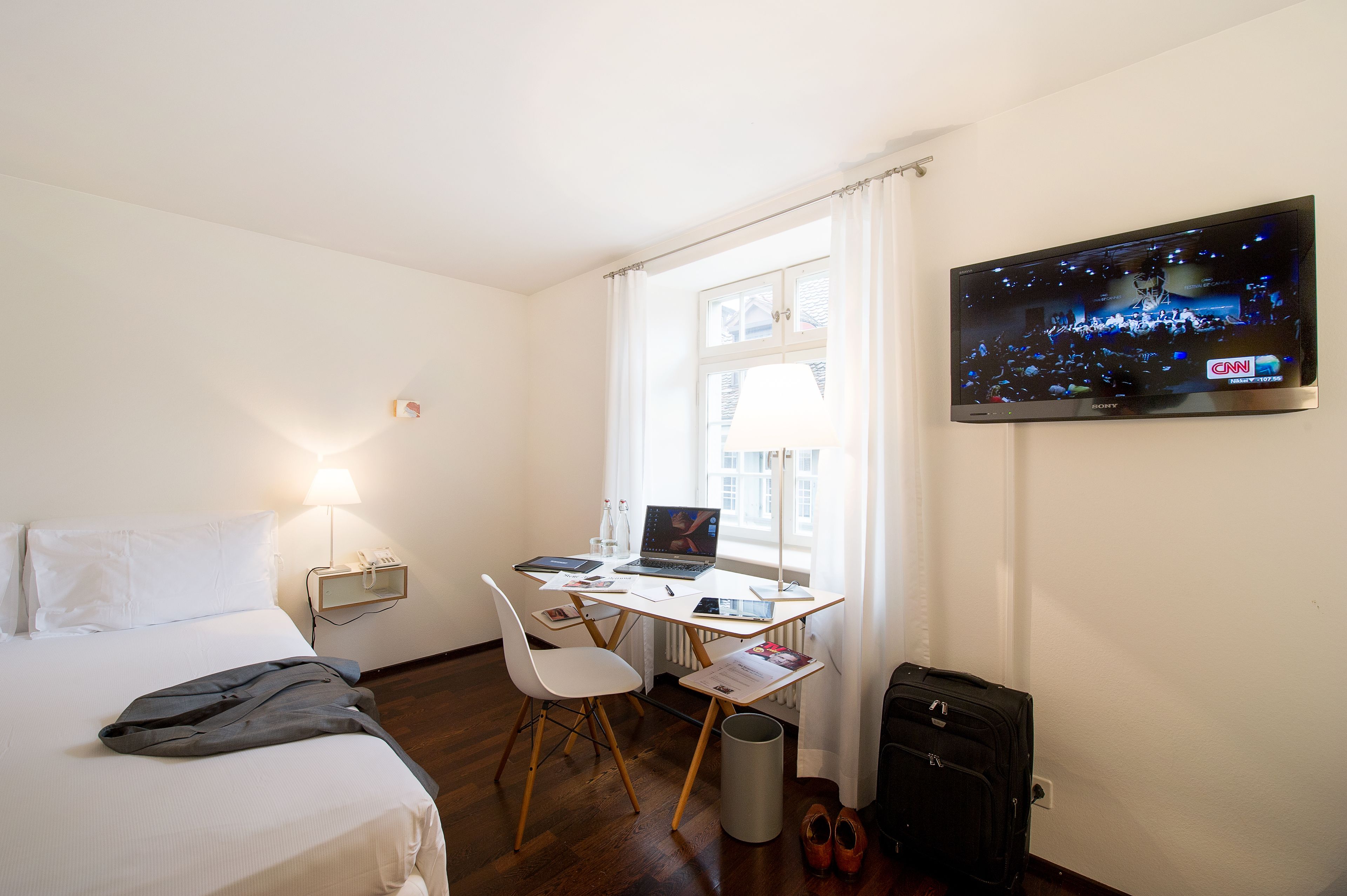A hotel room with a bed, a desk with a laptop, a TV on the wall, and a suitcase.
