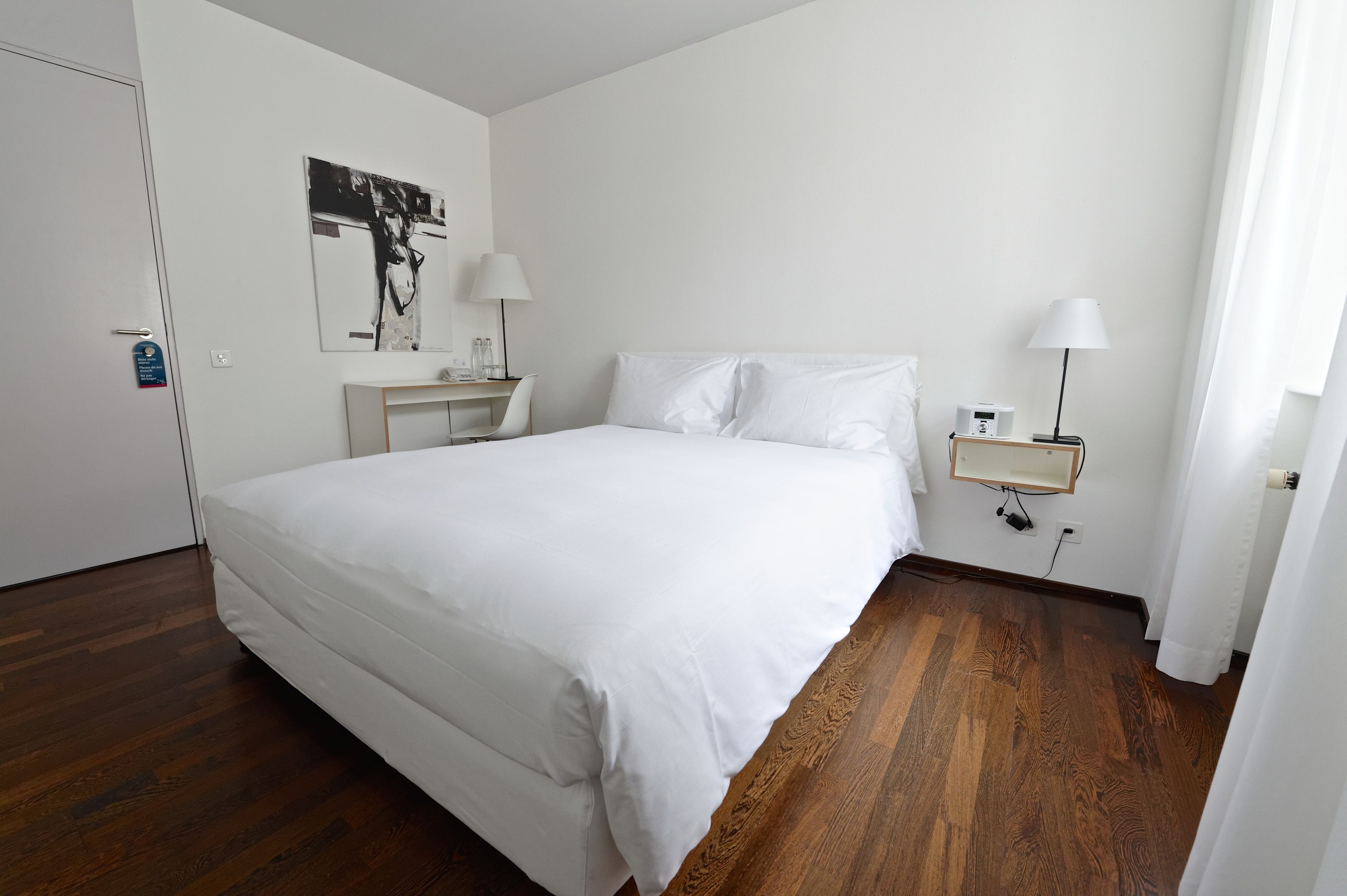 A hotel room with a large bed, a bedside table, a lamp, and a wall-mounted shelf.