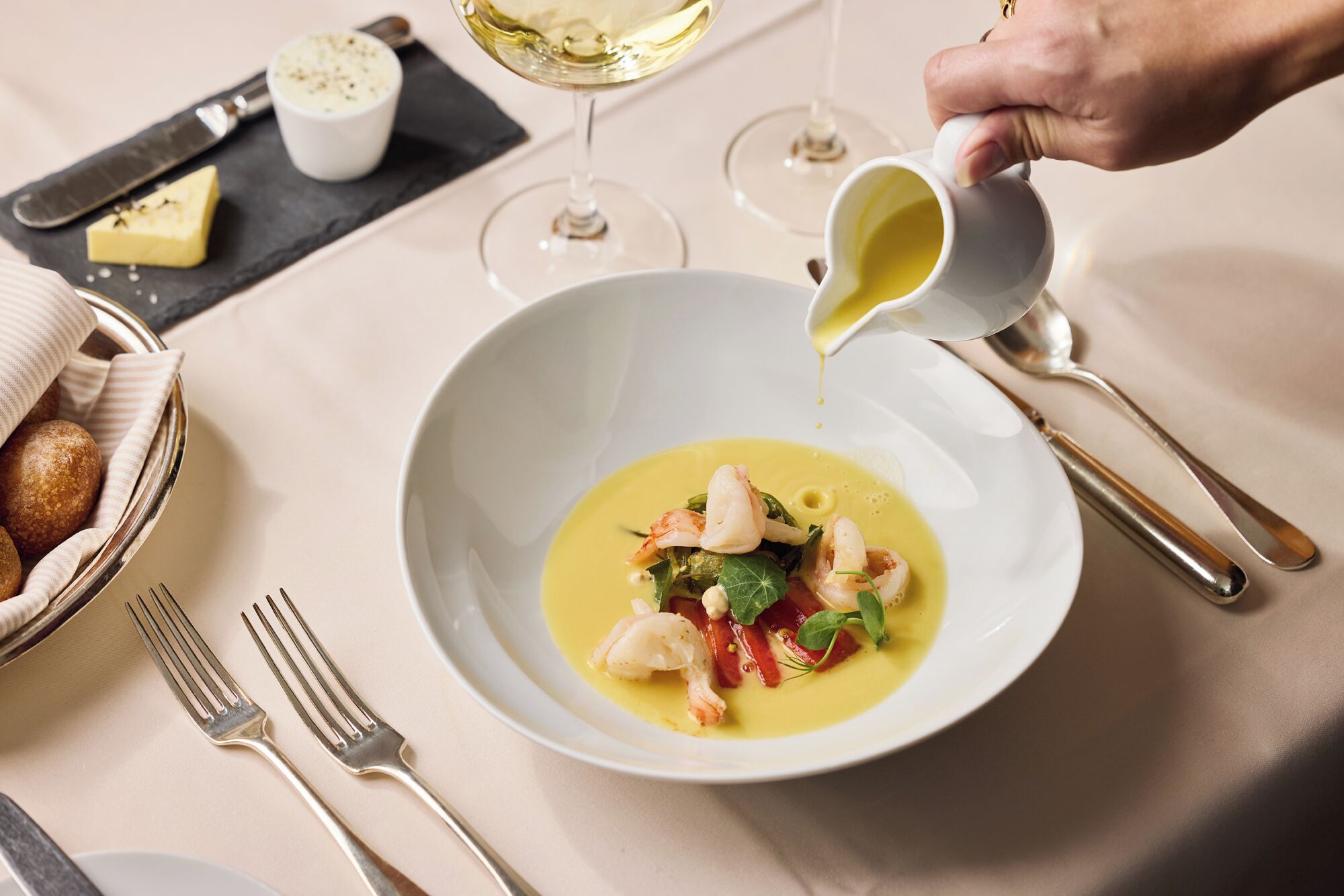 Una mesa de comedor con un plato de mariscos blancos, un bol de pan y una copa de vino blanco.