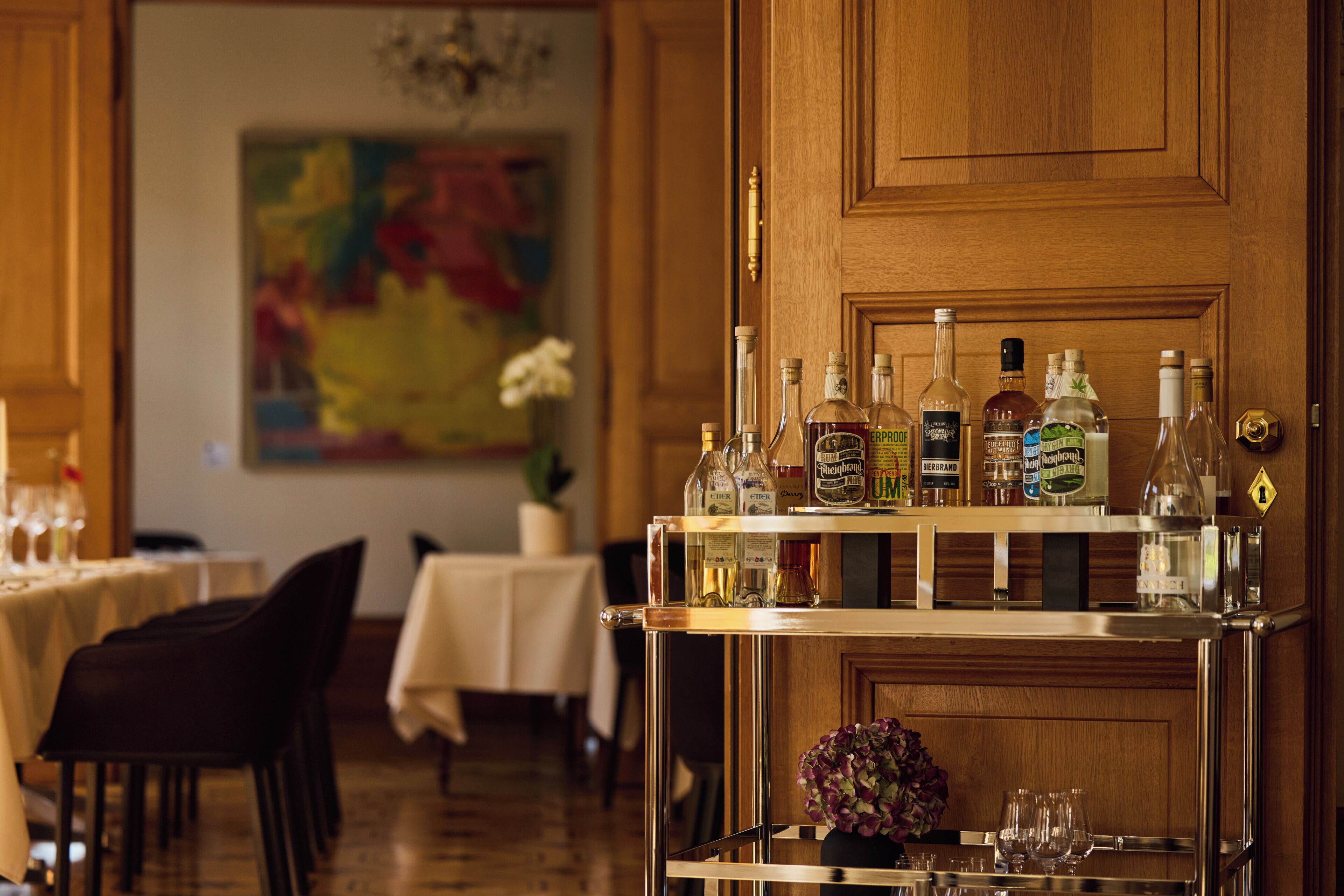 A bar area with a wooden door, a cart with bottles, and tables with chairs in the background.
