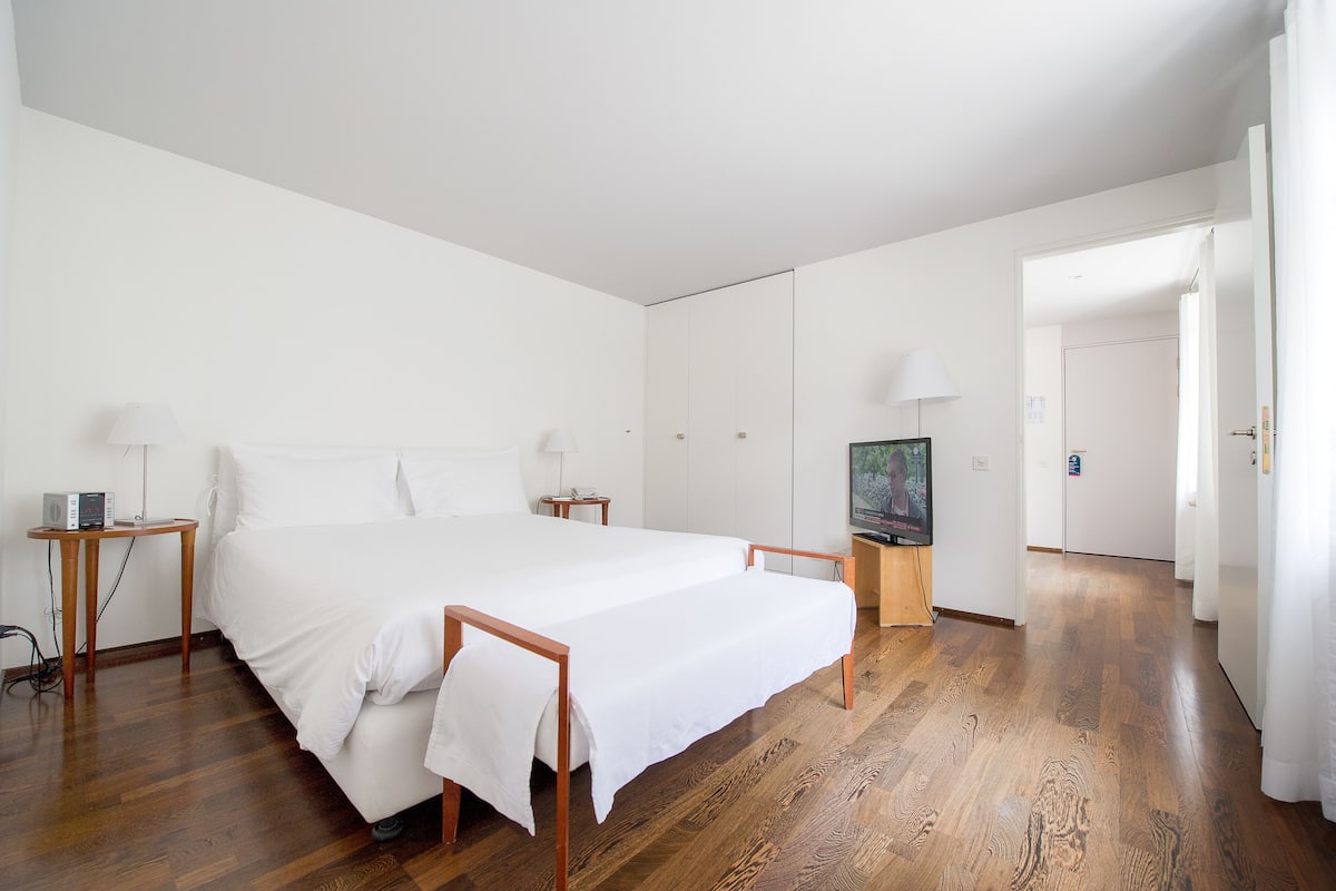 Un dormitorio espacioso con una cama grande, suelo de madera, paredes blancas y un televisor sobre un mueble de madera.