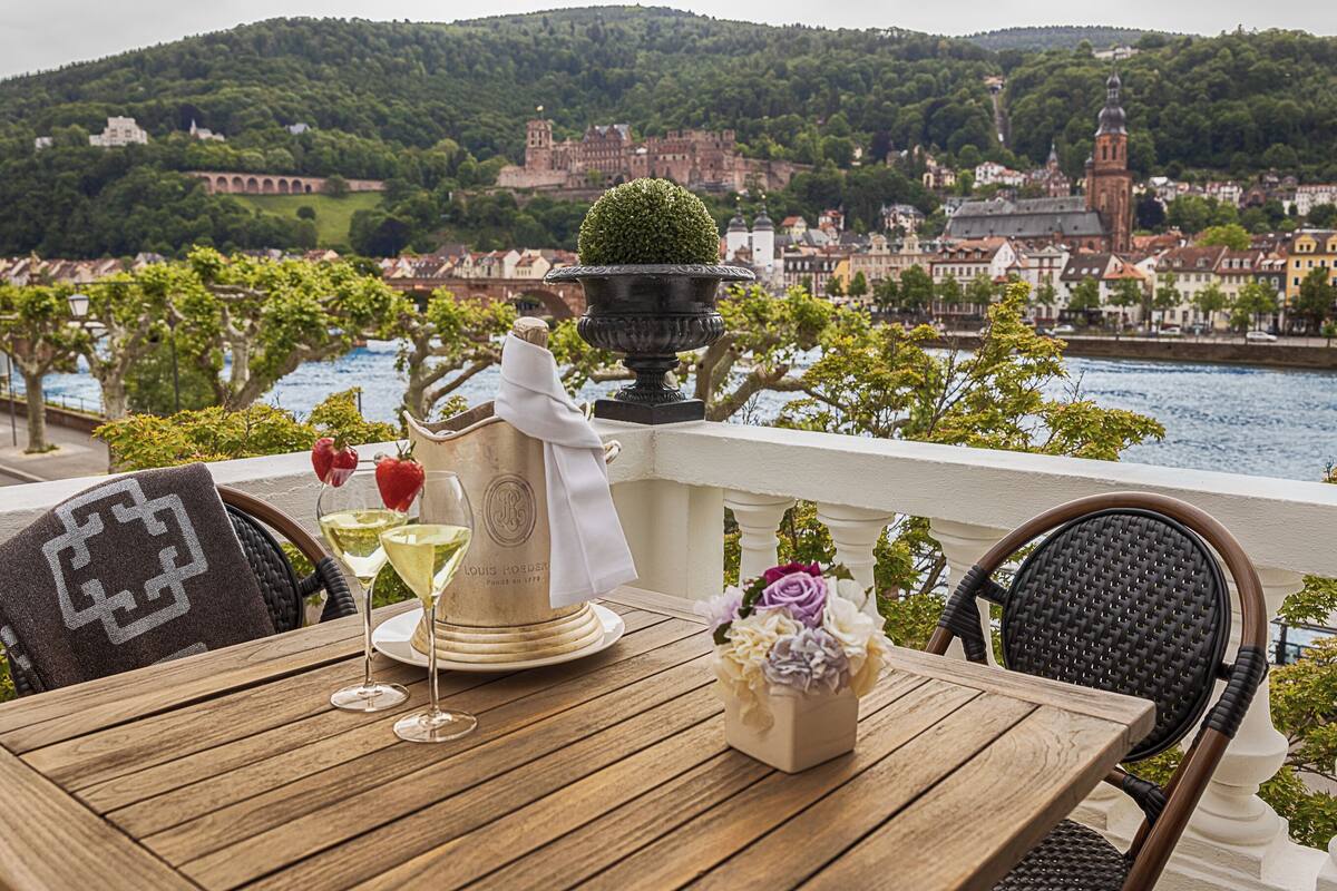A table set with a bottle, glasses, and flowers, overlooking a river and historic buildings.