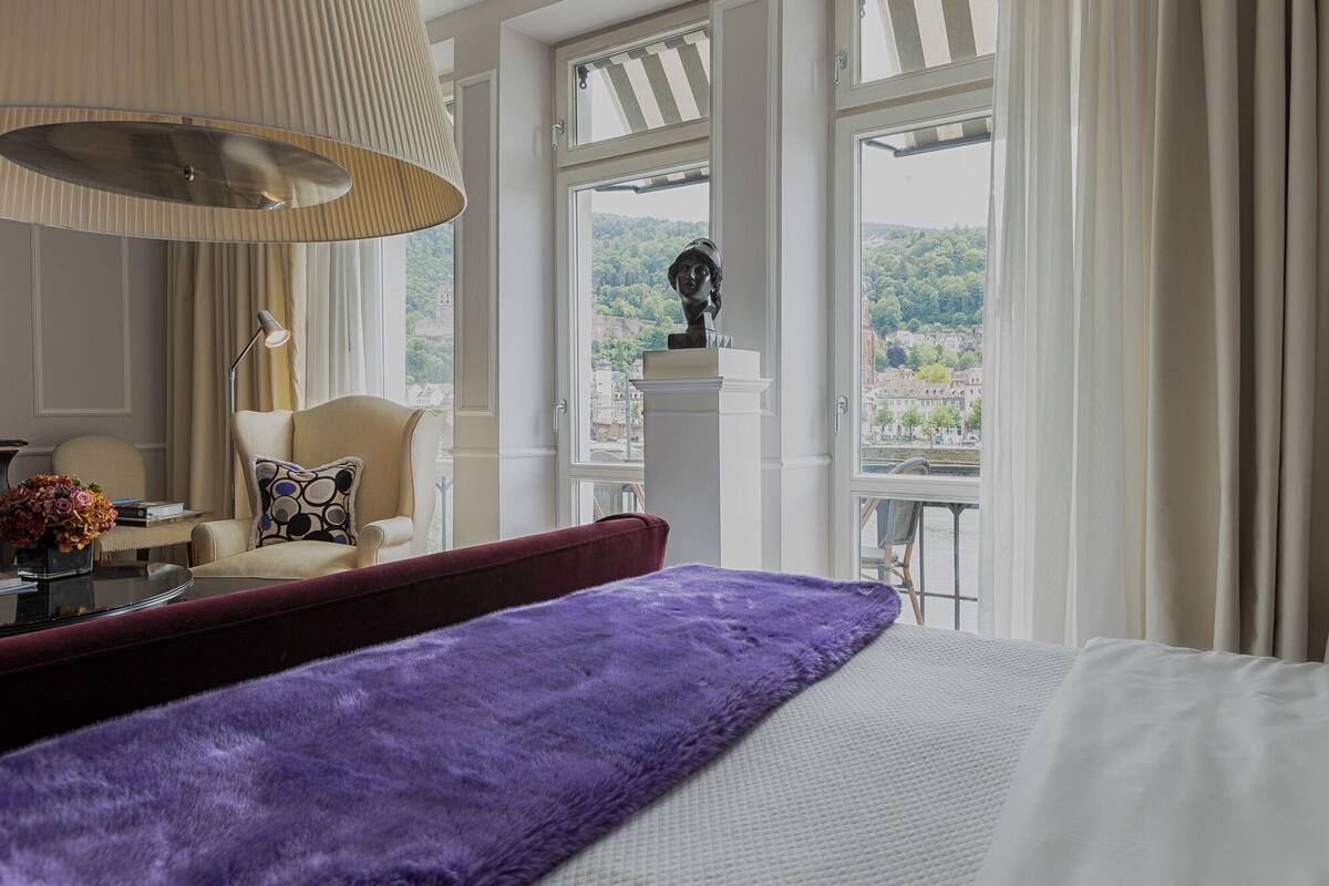 A hotel room with a large bed, a chair, a desk with a lamp, and a view of a cityscape through large windows.