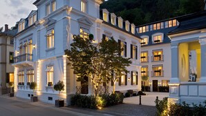 Een groot, meerlaags hotel in klassieke Europese stijl, met symmetrische ramen en een centrale binnenplaats met bomen en een bankje.