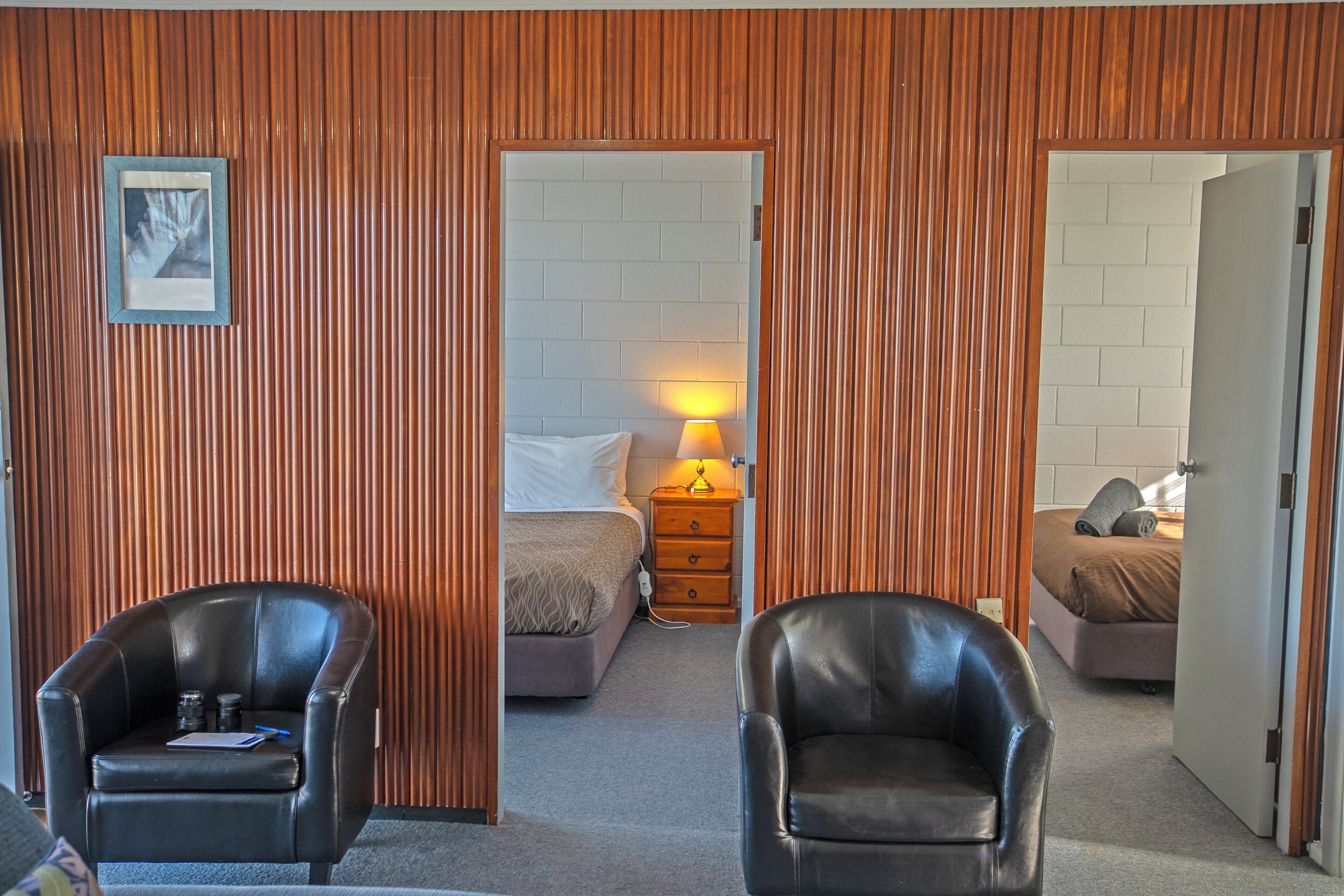 A room with two black leather chairs, a bed with a white headboard, a nightstand with a lamp, and a wooden slatted wall.