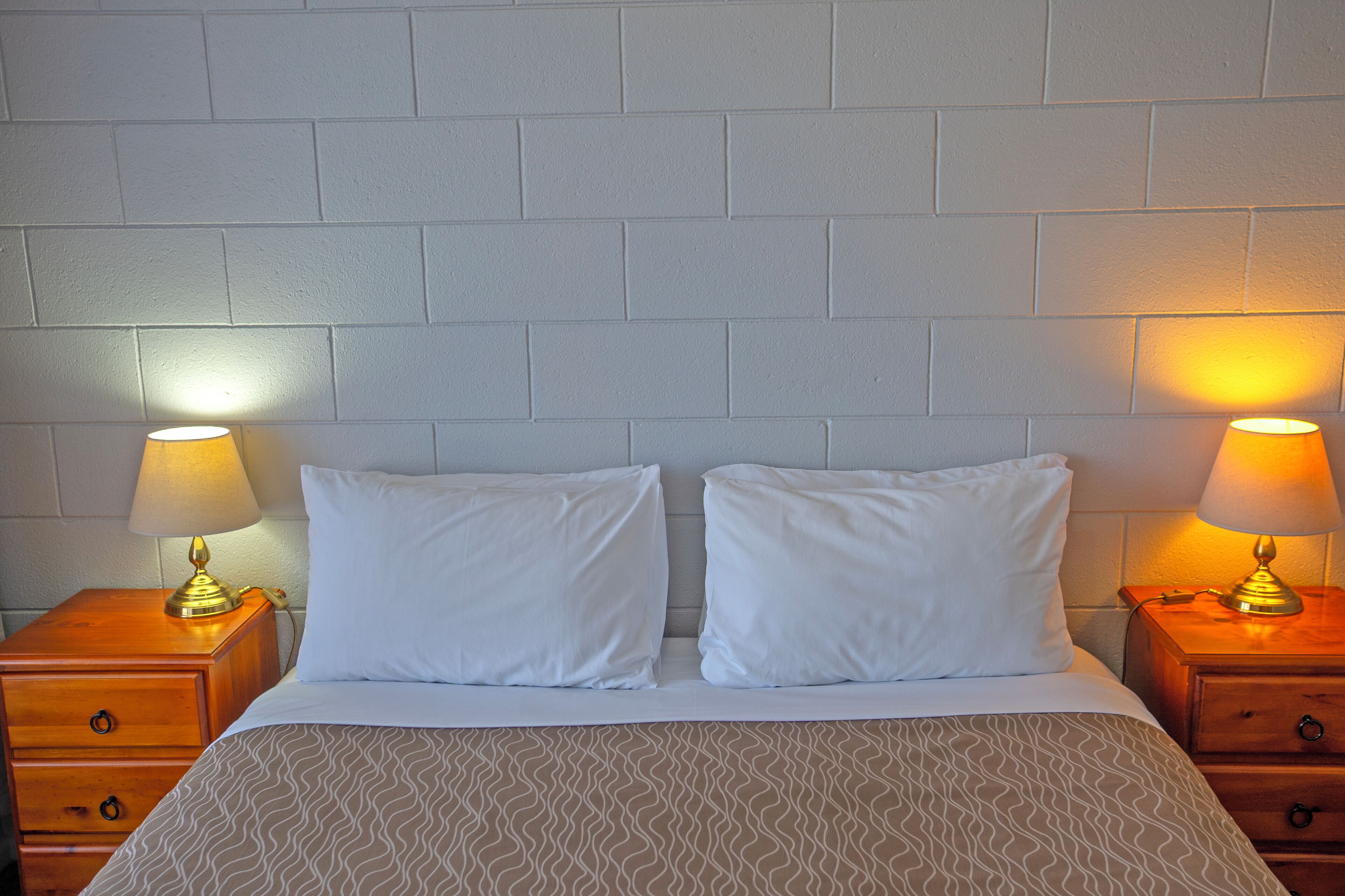 A neatly made bed with two white pillows and a patterned bedspread, flanked by two bedside tables with lamps, against a plain white brick wall.