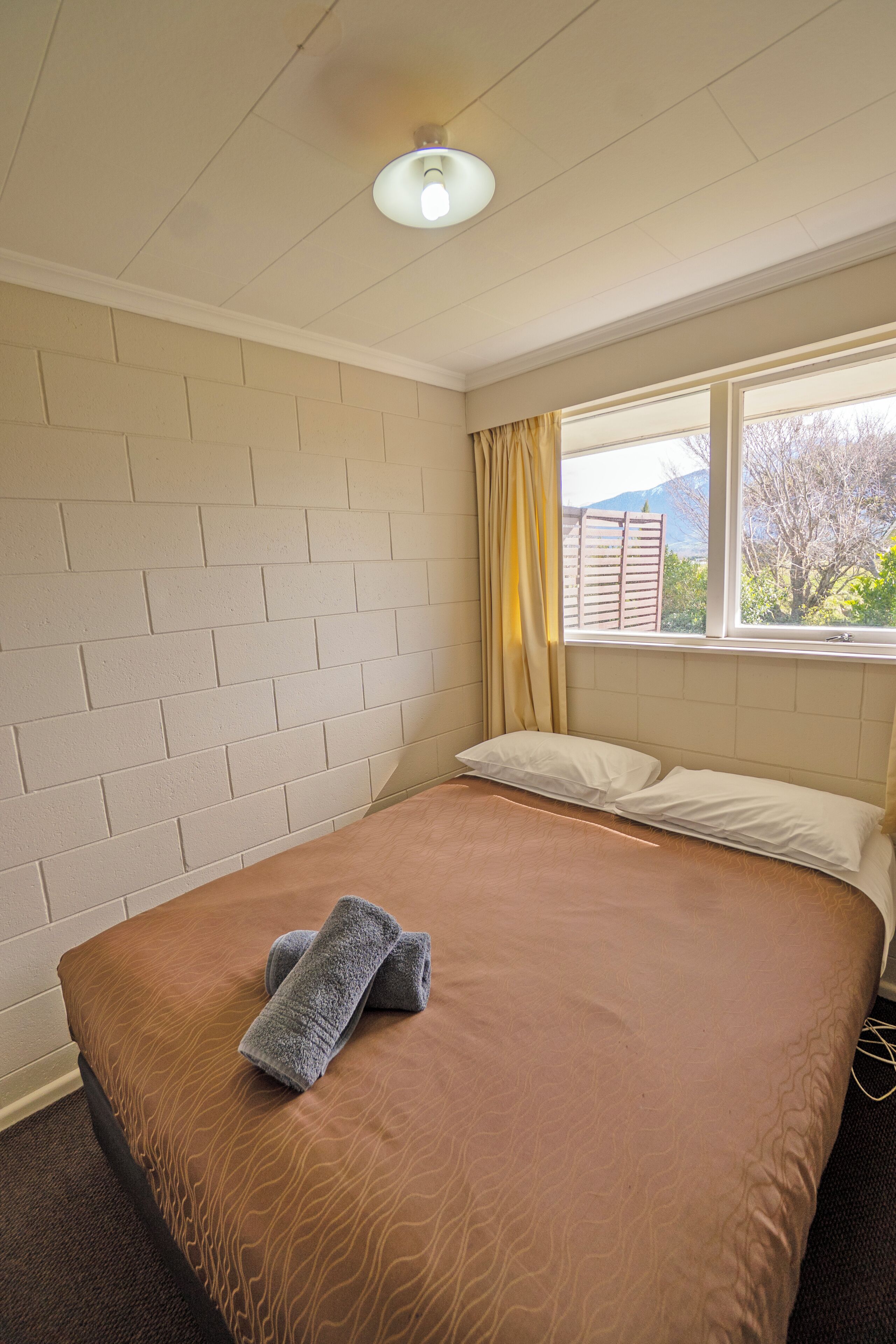 A neatly made bed with a brown patterned bedspread and two rolled towels, a window with curtains, and a brick-patterned wall.