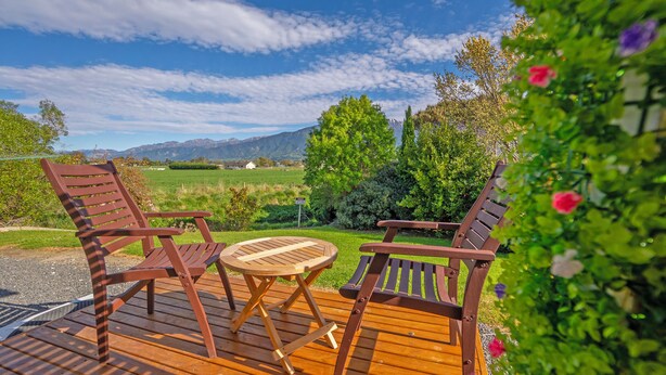 A wooden deck with two chairs and a small round table, overlooking a green landscape with trees and mountains.