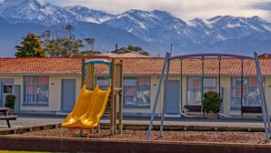 A playground with a yellow slide, swings, and a building with a red-tiled roof in the background.
