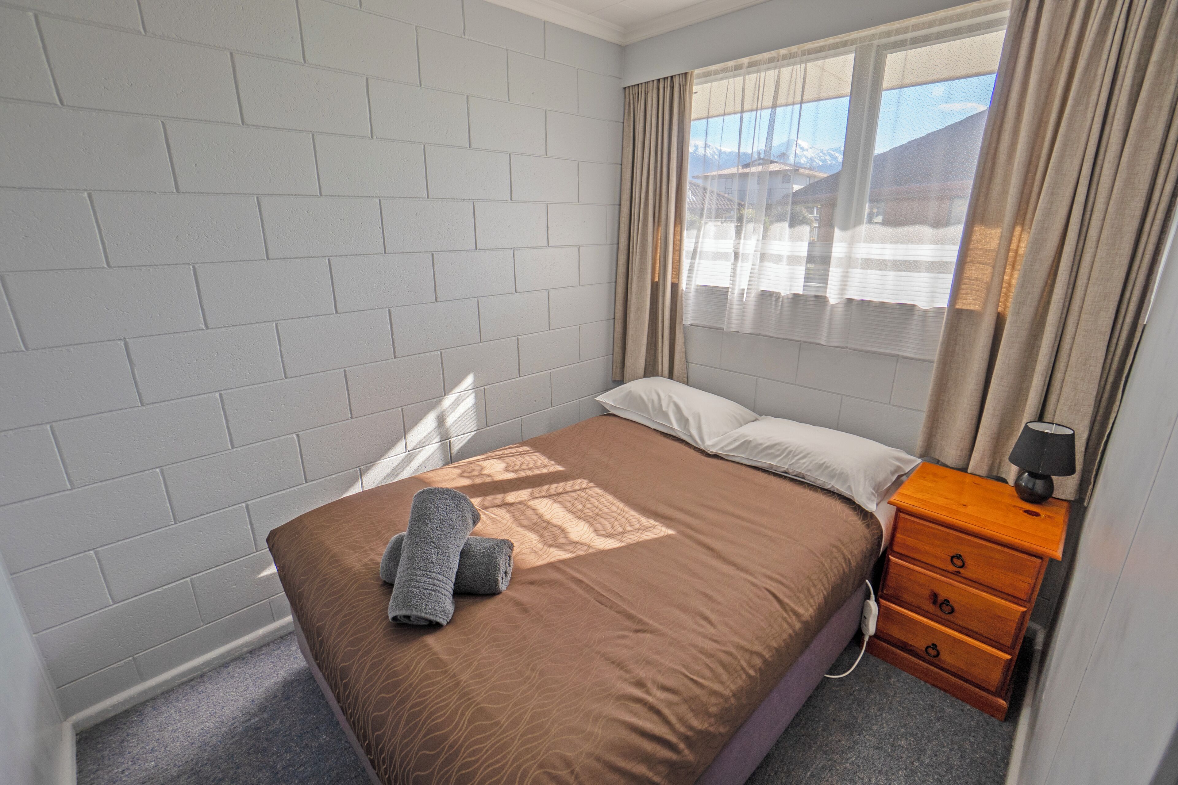 A hotel room with a bed, a nightstand, a window with curtains, and a brick wall.