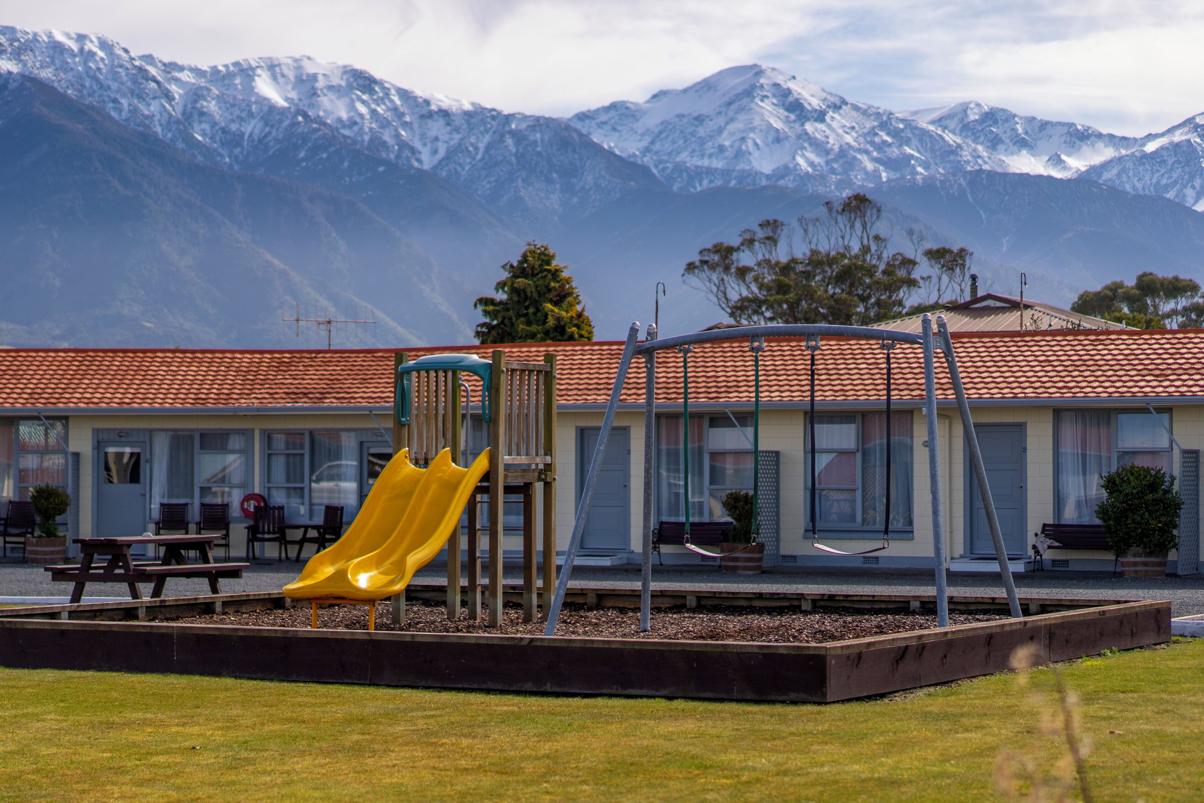 A playground with a yellow slide, picnic tables, and a building with a red-tiled roof.