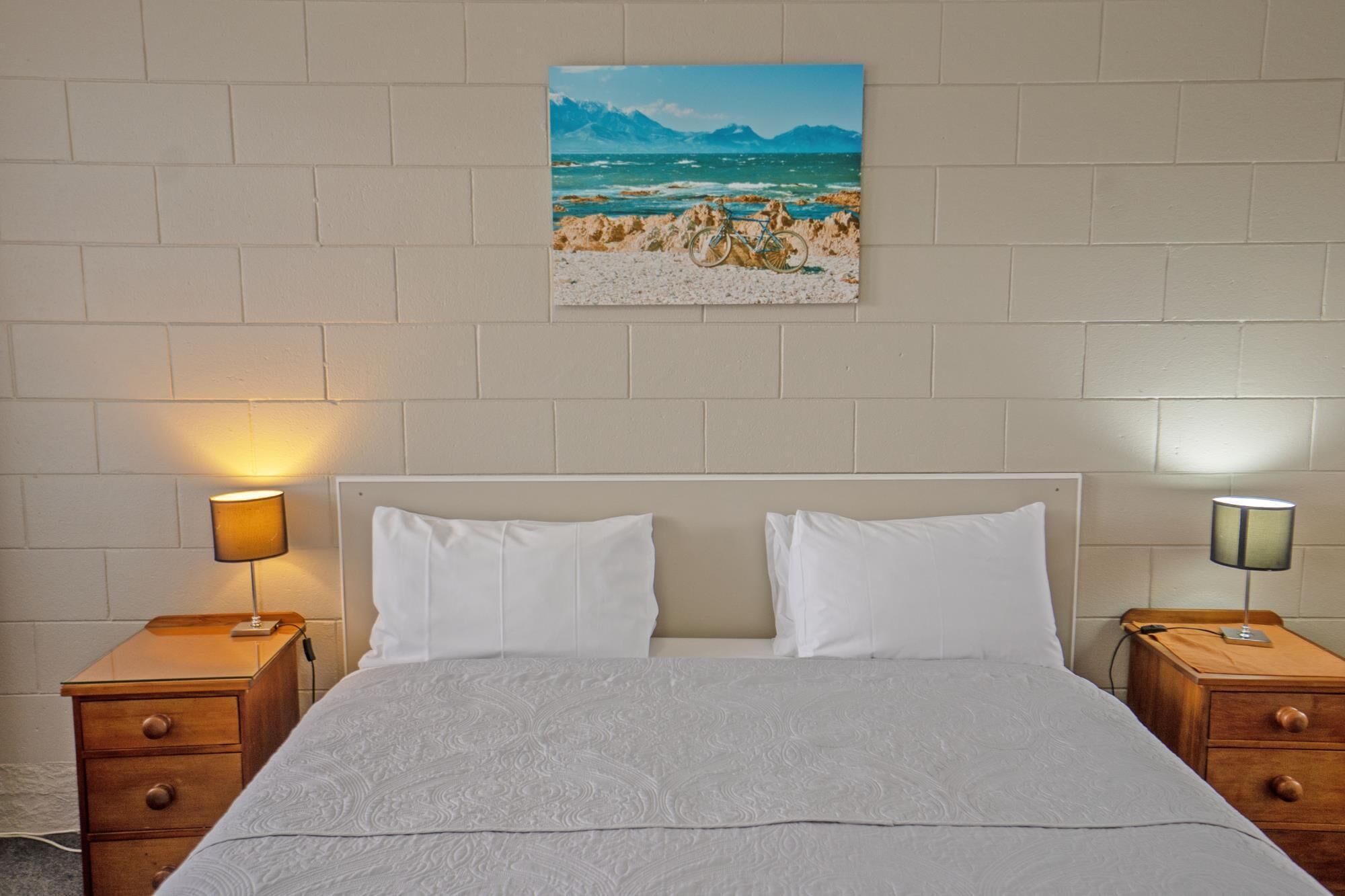 Un dormitorio con una cama, dos mesitas de noche, una lámpara y un cuadro de una playa.